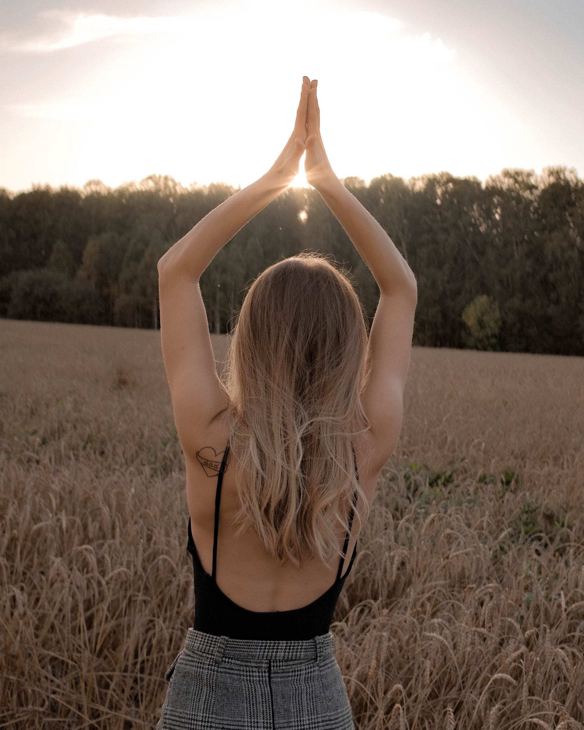 A woman with blond hair stands in a wheat field with hands folded in prayer at sunset.